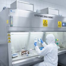 Stainless steel cytotoxic drug cabinet undergoing containment validation while a technician handles IV bags in a sterile cleanroom