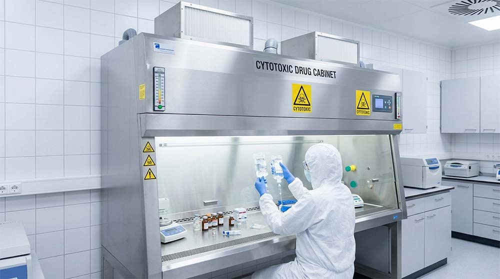 Stainless steel cytotoxic drug cabinet undergoing containment validation while a technician handles IV bags in a sterile cleanroom